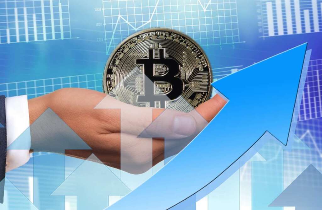 una grossa moneta di Bitcoin nel palmo di una mano. Nello sfondo dei grafici finanziari, e alcune frecce verso l'alto in sovrimpressione